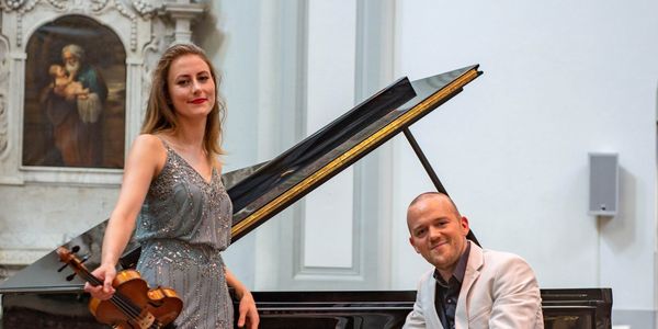A woman in a silver dress holds a violin beside a man in a white suit seated at a grand piano in an elegant room with a decorative wall.