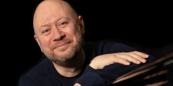 Black and white image of a person in a suit holding a saxophone, leaning against a wall with a music stand nearby.