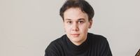 Marek Kąkol in a black turtleneck leans on a grand piano, looking at the camera against a plain background.