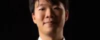 A person in a black suit with a white shirt and tie, smiling against a dark background.