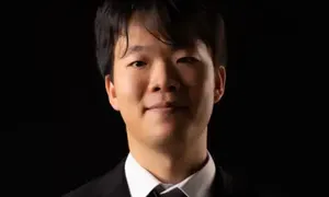 A person in a black suit with a white shirt and tie, smiling against a dark background.
