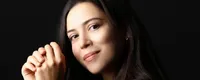 Woman with long dark hair in a black shirt, sitting with hands clasped, poses against a black background, looking at the camera.