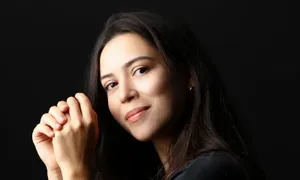 Woman with long dark hair in a black shirt, sitting with hands clasped, poses against a black background, looking at the camera.