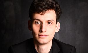 Young man in a black suit and shirt, posing with crossed arms on a white furry surface against a dark background.