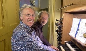 Two people playing an organ, one smiling at the camera. Music sheets are visible on the organ.