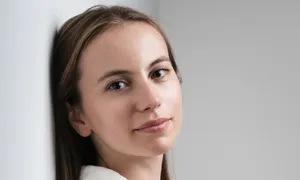 Woman with long hair in a white shirt, leaning against a wall, looking at the camera with a soft smile. Neutral background.