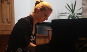 Woman playing a grand piano in a room with wooden paneling and a potted plant in the background.