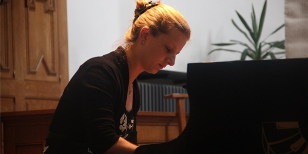 Woman playing a grand piano in a room with wooden paneling and a potted plant in the background.