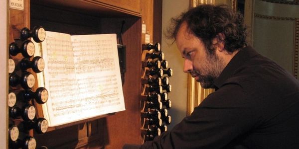Smiling person wearing glasses and a colorful shirt, standing in front of an organ.