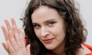 A woman with curly hair in a red sweater, smiling and holding her hands together in a soft, natural light setting.