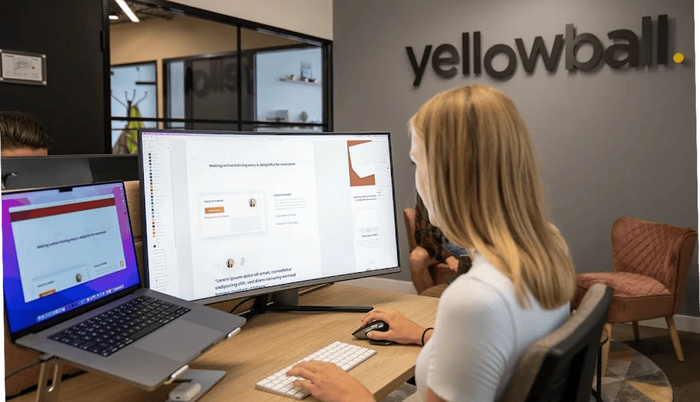 Woman working at a desk with dual monitors in an office with "yellowball" logo on the wall.