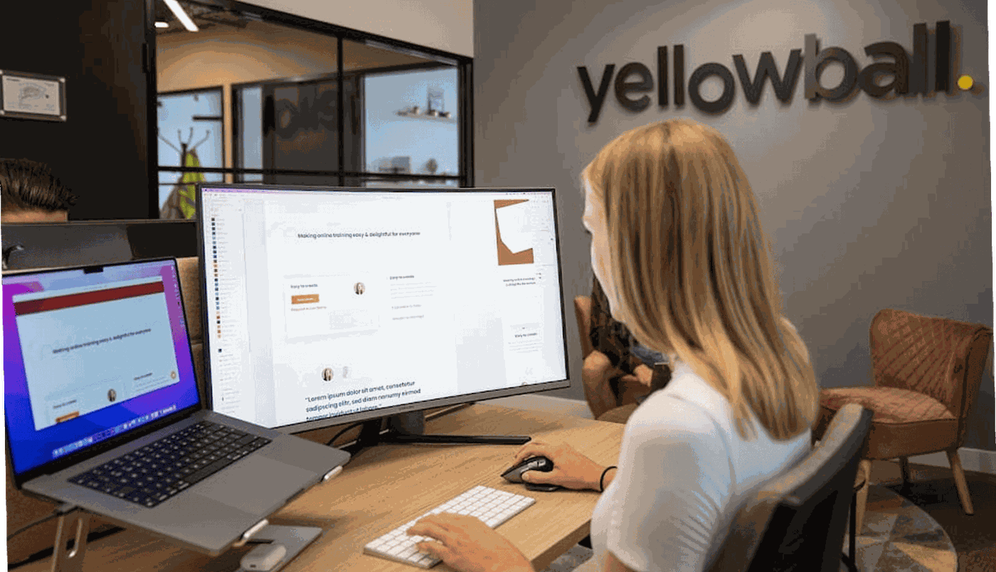 Woman working at a desk with dual monitors in an office with "yellowball" logo on the wall.