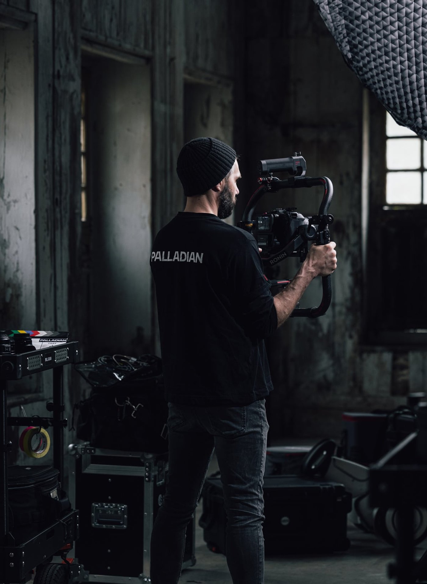 Man in a beanie and "PALLADIAN" shirt operates a camera stabilizer in a dimly lit, rustic room with film equipment around.