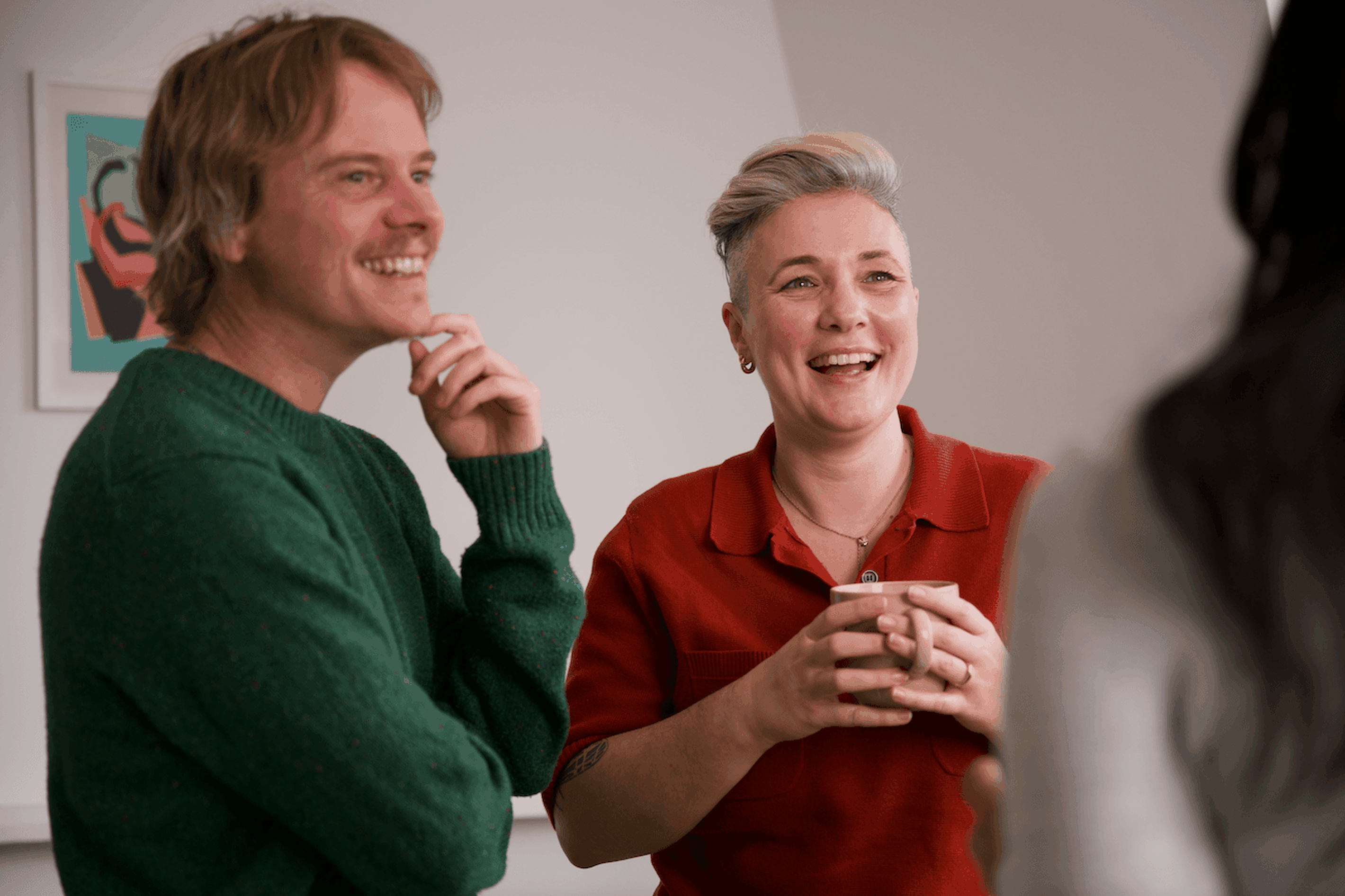 Two people smiling and chatting indoors, one holding a mug. A framed artwork is visible on the wall behind them.