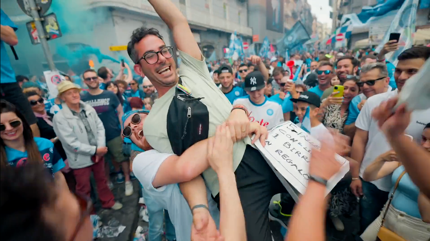 A joyful crowd celebrates, lifting a smiling man in the air. People around hold flags and signs amidst a festive street scene.