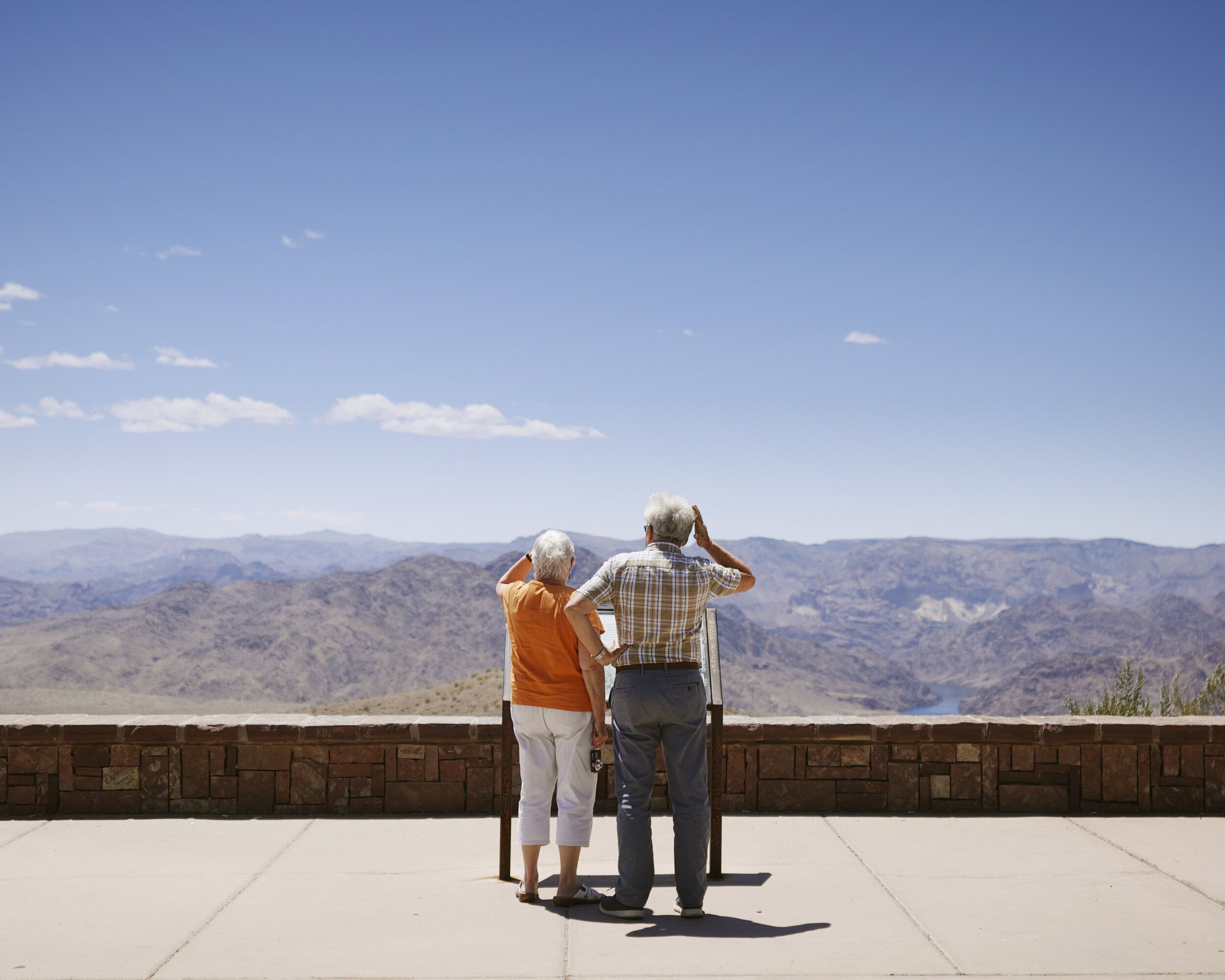 An elderly couple stands on a viewing platform, admiring a vast mountain landscape under a clear blue sky.