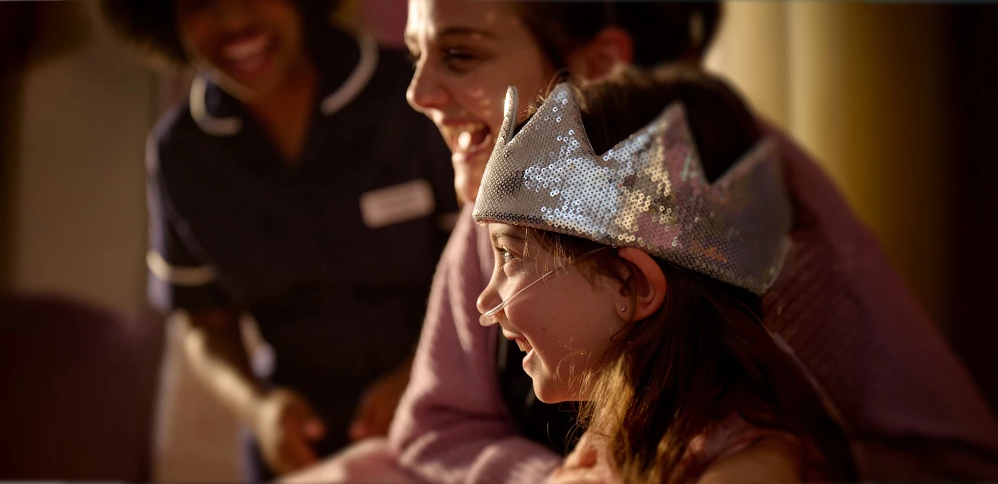 Child wearing a silver crown smiles alongside an adult as a nurse stands nearby, creating a joyful and warm atmosphere.