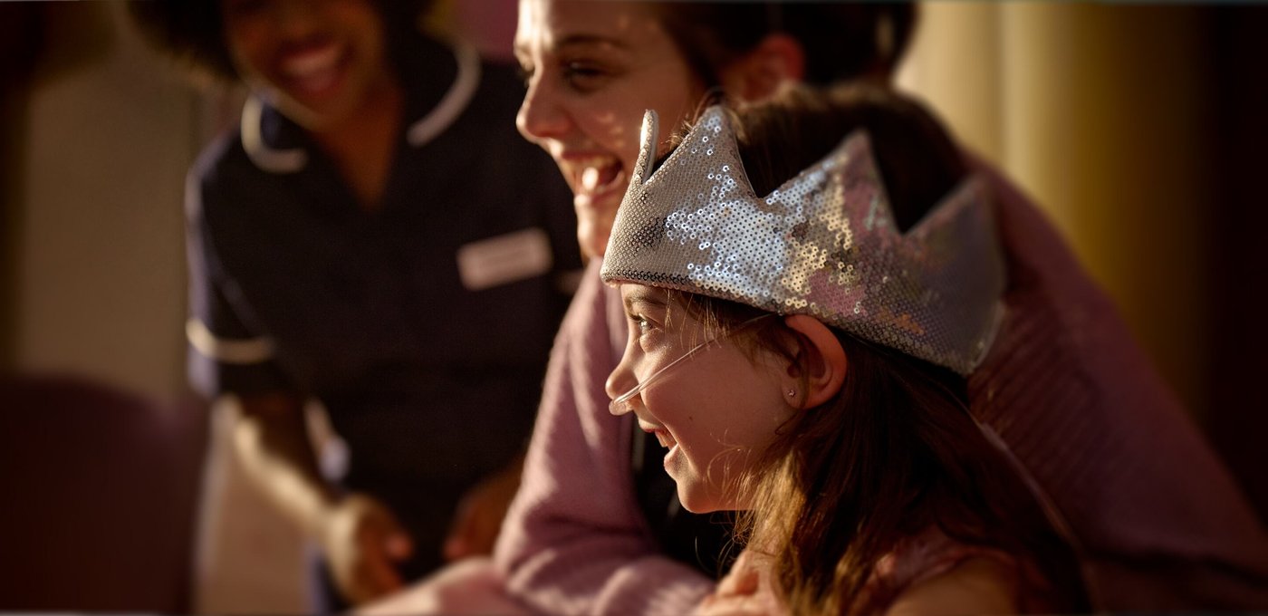 Child wearing a silver crown smiles alongside an adult as a nurse stands nearby, creating a joyful and warm atmosphere.