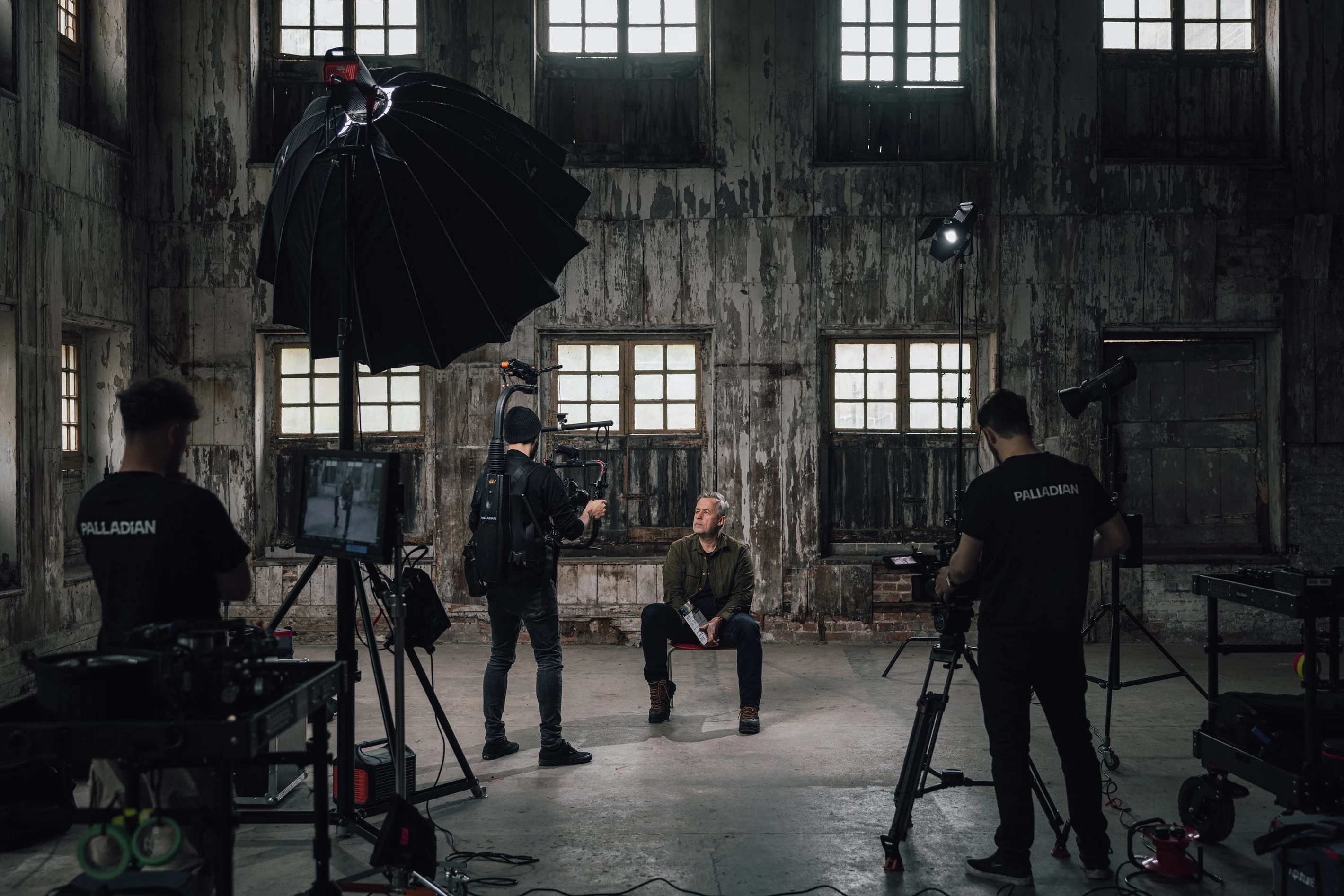 Film crew setting up to shoot a scene with a seated actor in a dimly lit, rustic room with multiple windows and professional lighting equipment.