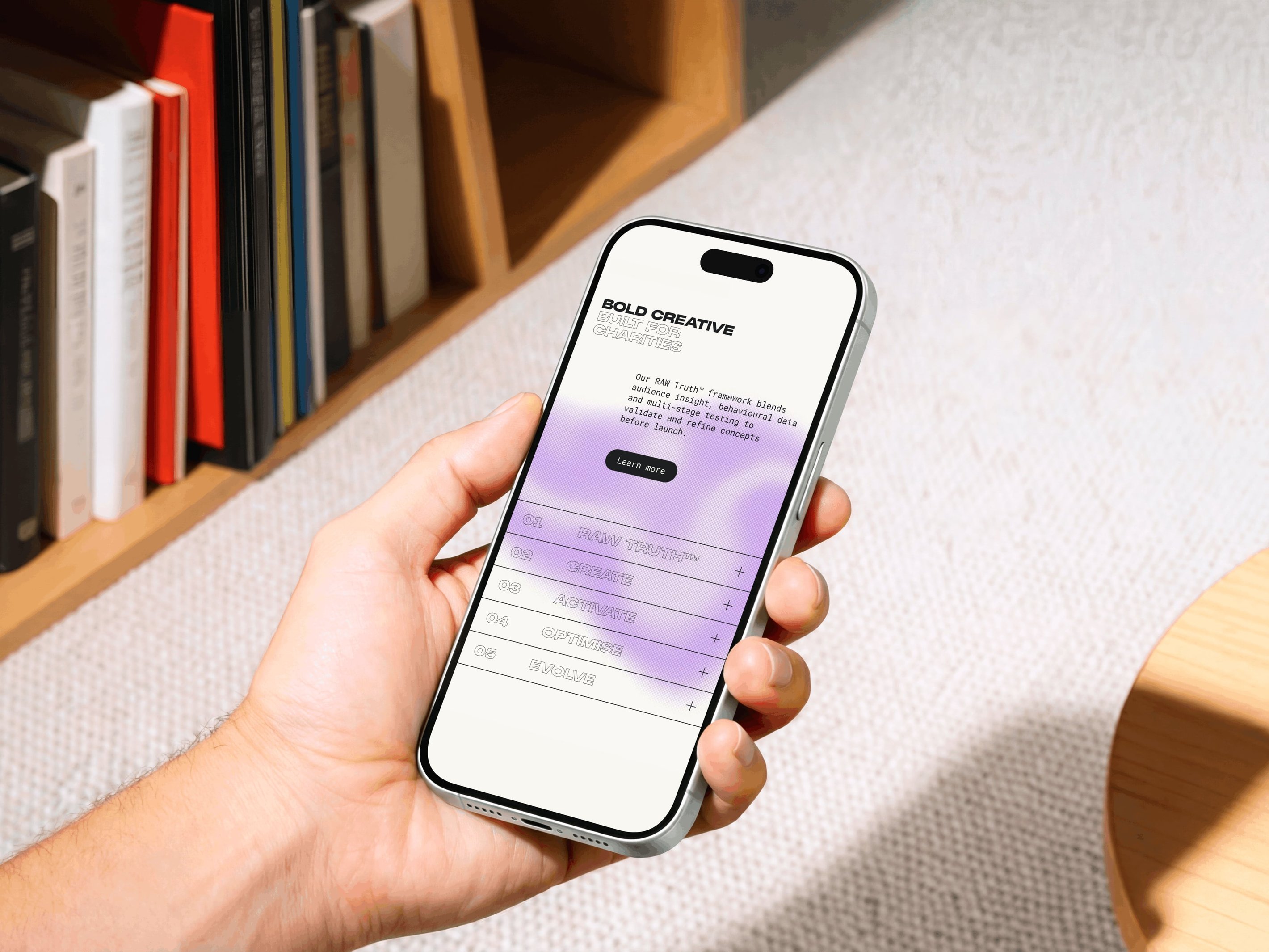 Hand holding a smartphone displaying a website with the title "Bold Creative" and pastel purple gradient. Bookshelf in the background.