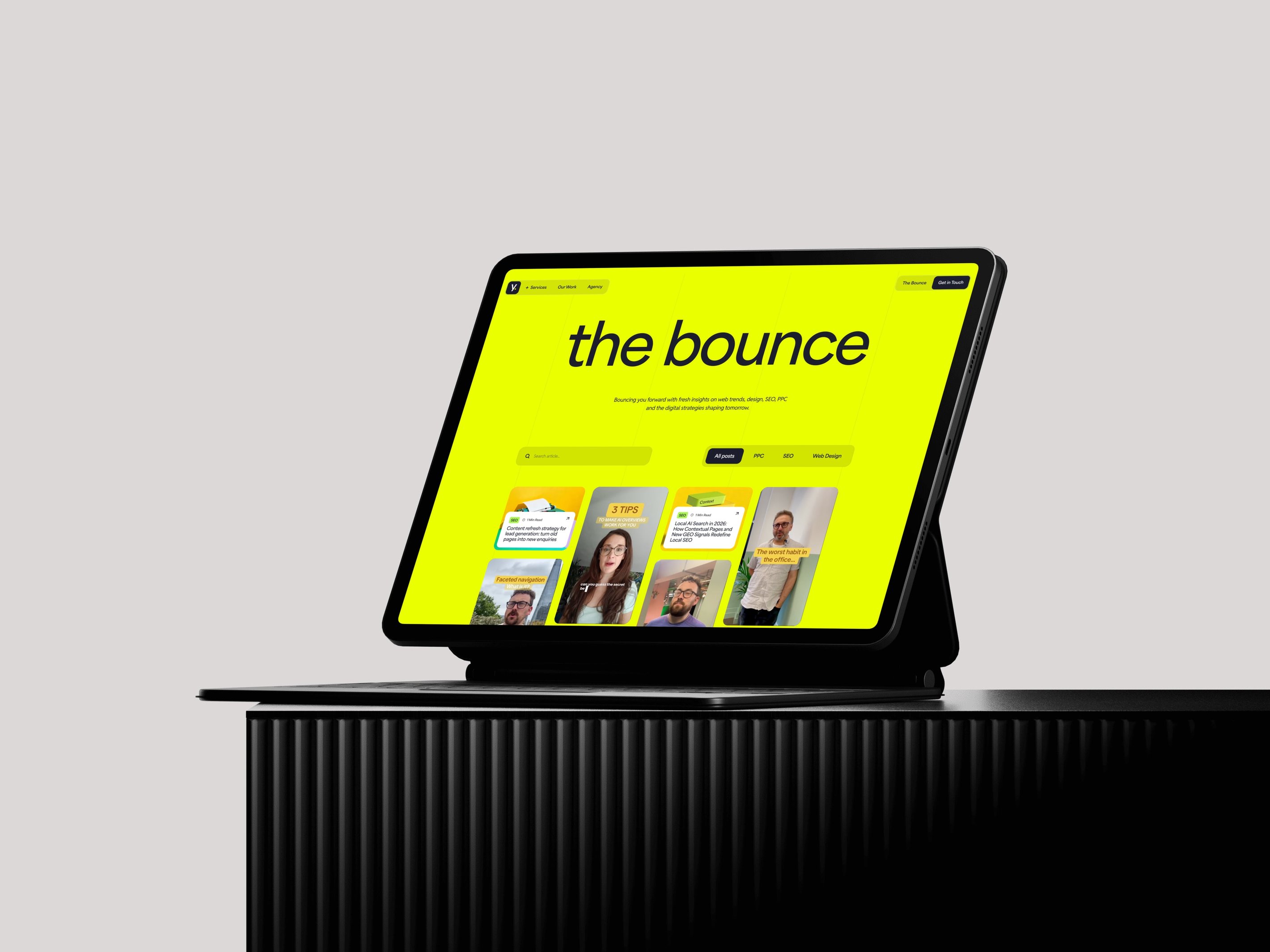 Tablet on a dark surface displaying a bright yellow screen with "the bounce" text and various people images on it.