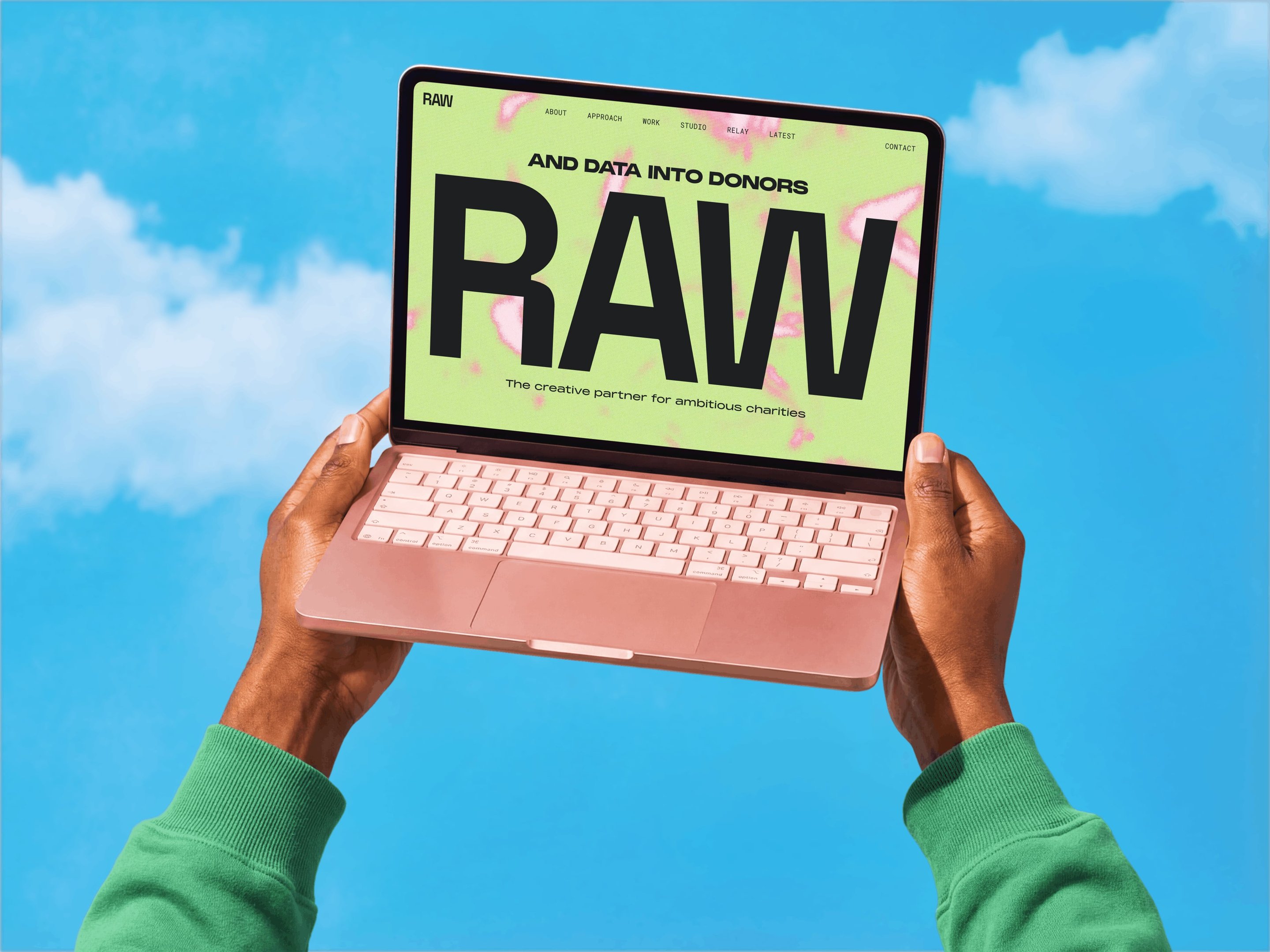 Hands holding a pink laptop against a blue sky, displaying a webpage with "RAW" in bold text and additional smaller text visible.