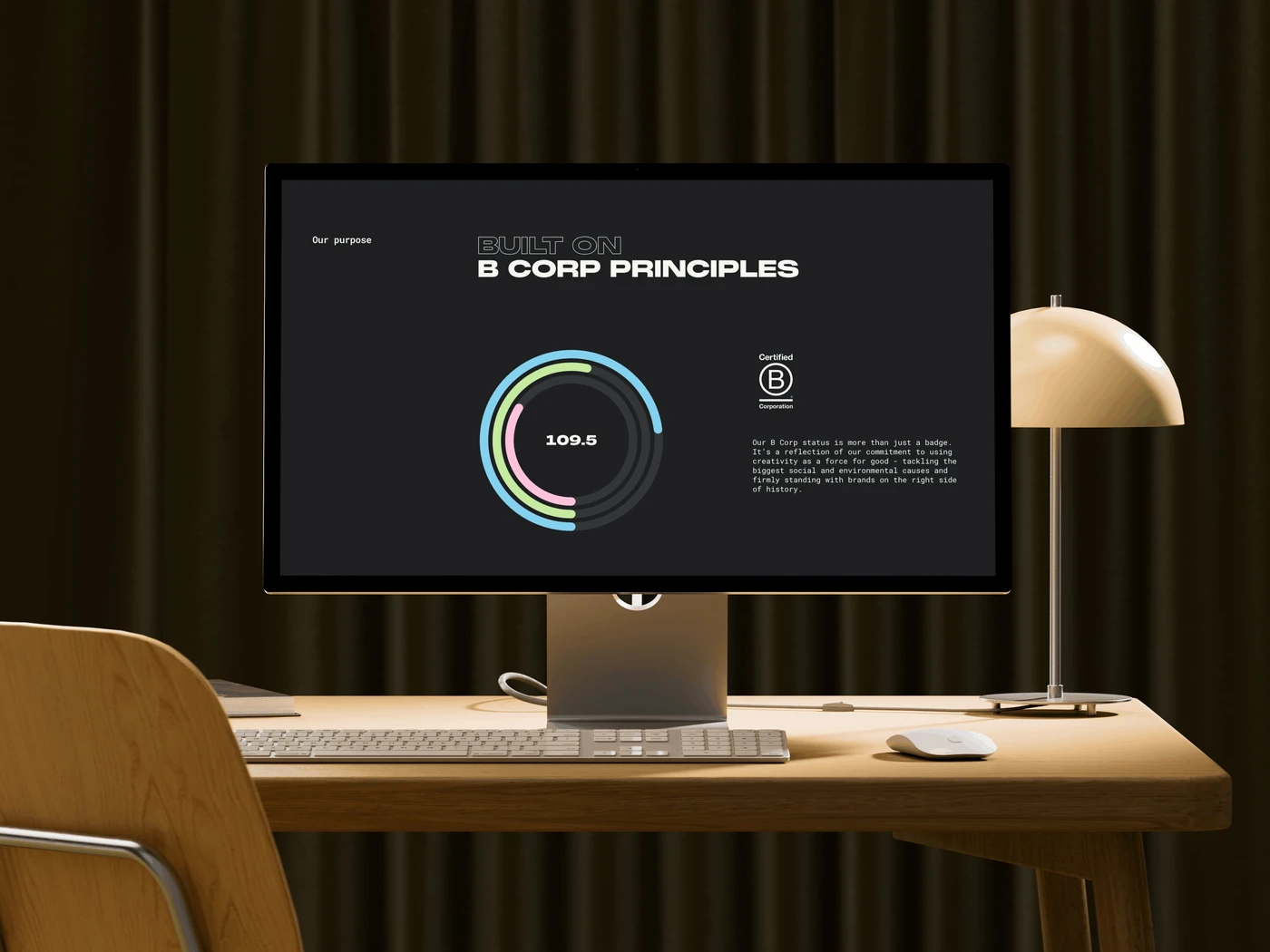 Computer screen showing "Built on B Corp Principles" with a score of 109.6. Desk lamp, keyboard, and mouse on a wooden desk in a dimly lit room.