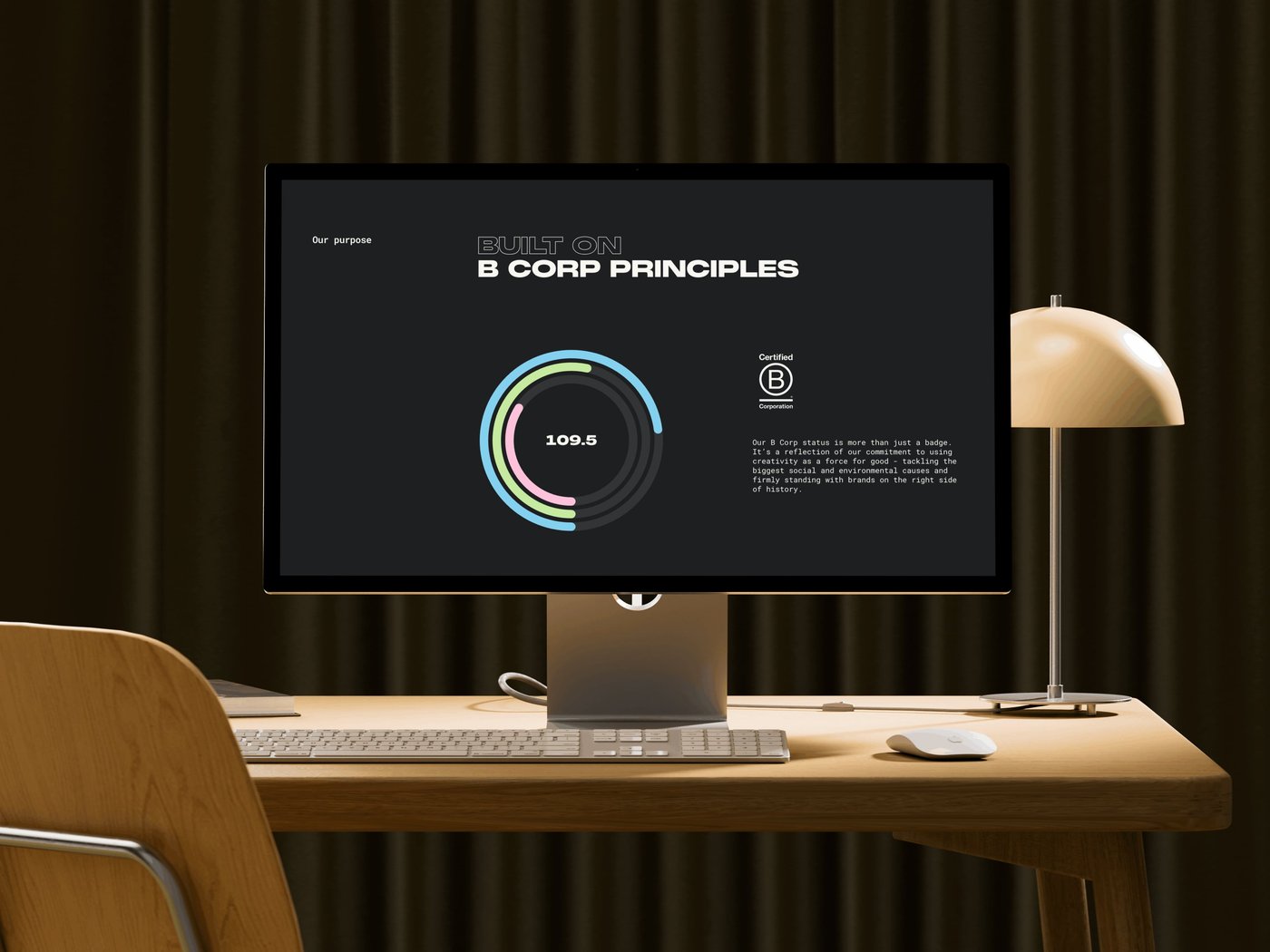Computer screen showing "Built on B Corp Principles" with a score of 109.6. Desk lamp, keyboard, and mouse on a wooden desk in a dimly lit room.