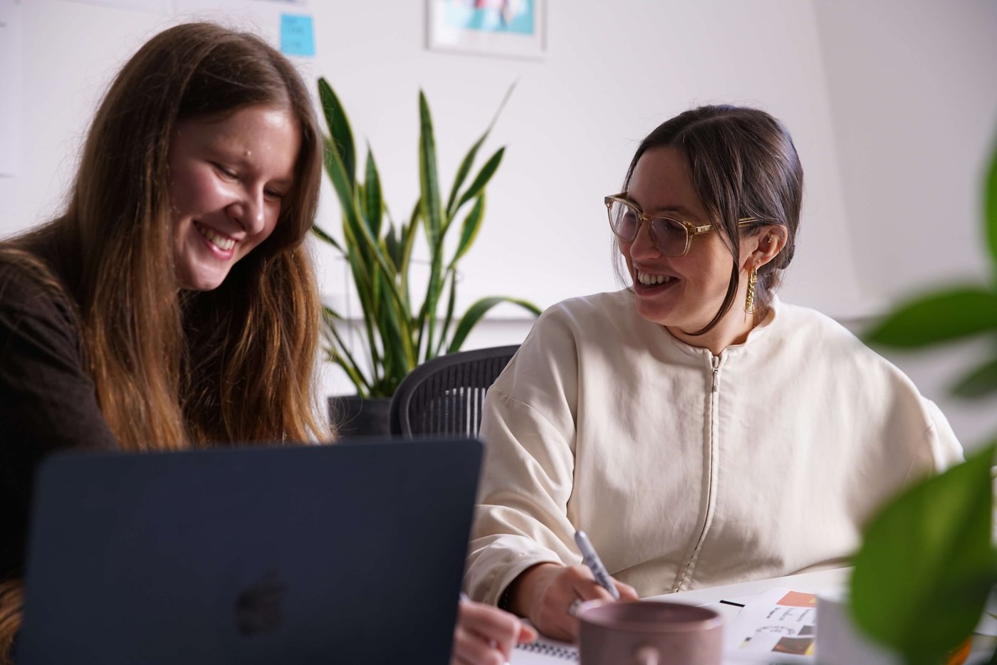 Two women smile and collaborate at a desk, one writing notes, the other using a laptop. Indoor plants and notes on the wall are in the background.