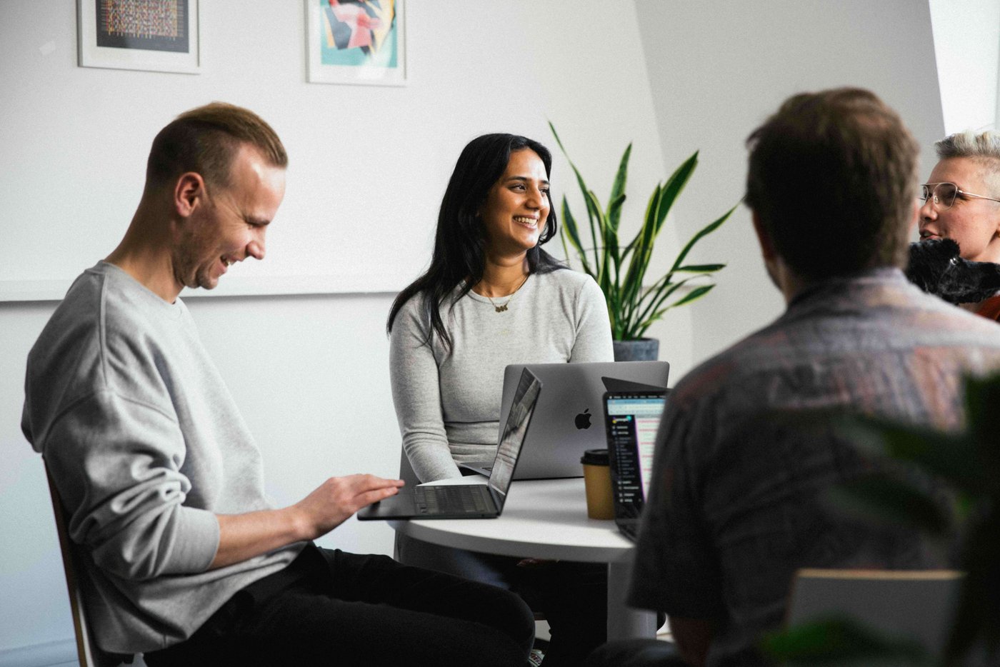 Four people in a casual meeting around a table with laptops, smiling and engaged in conversation, with potted plants in the background.