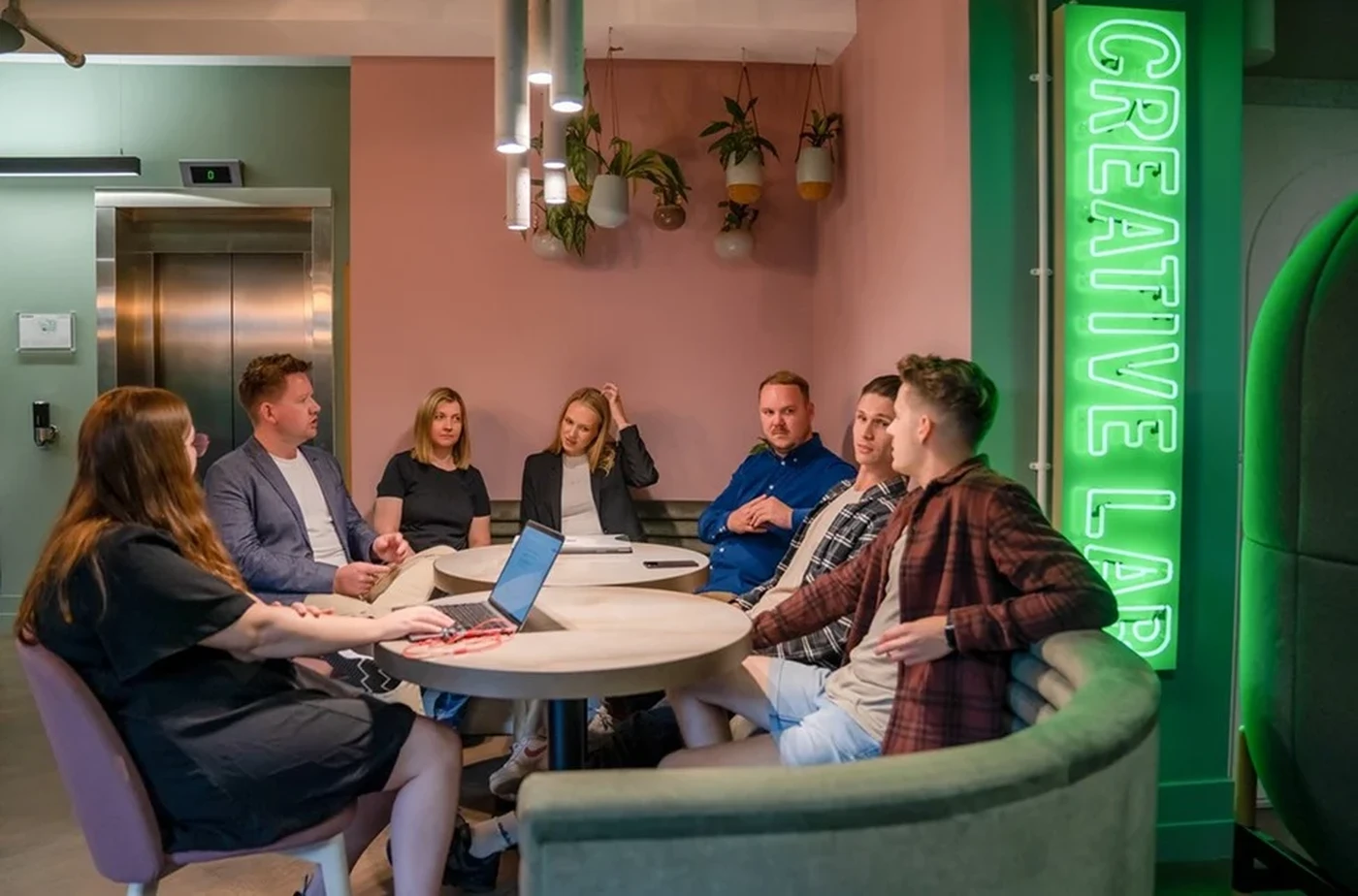 A group of people having a discussion in a modern meeting space with a neon "Creative Lab" sign and plants in the background.