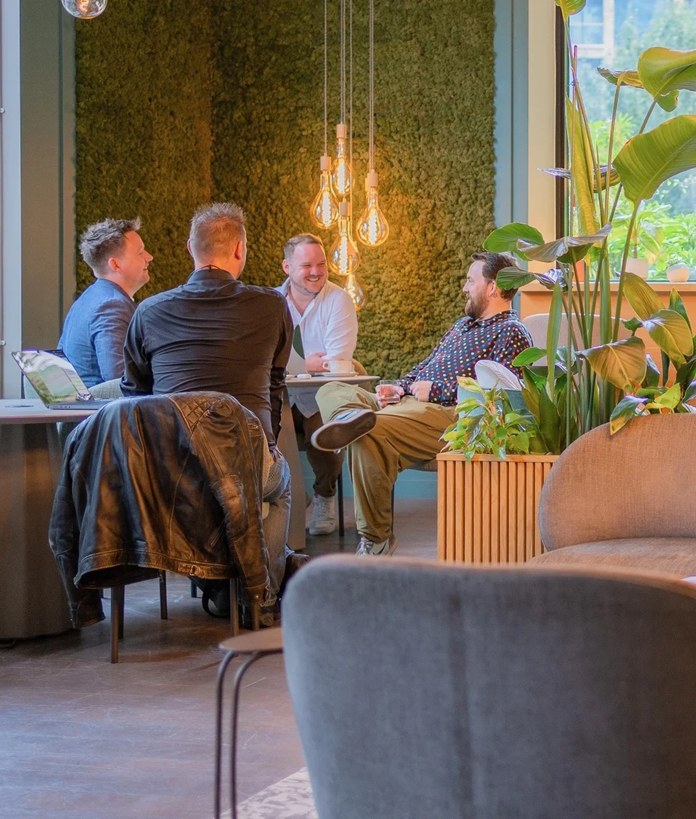 Four people sit around a table in a modern office with plants and hanging lights, engaged in a lively discussion.
