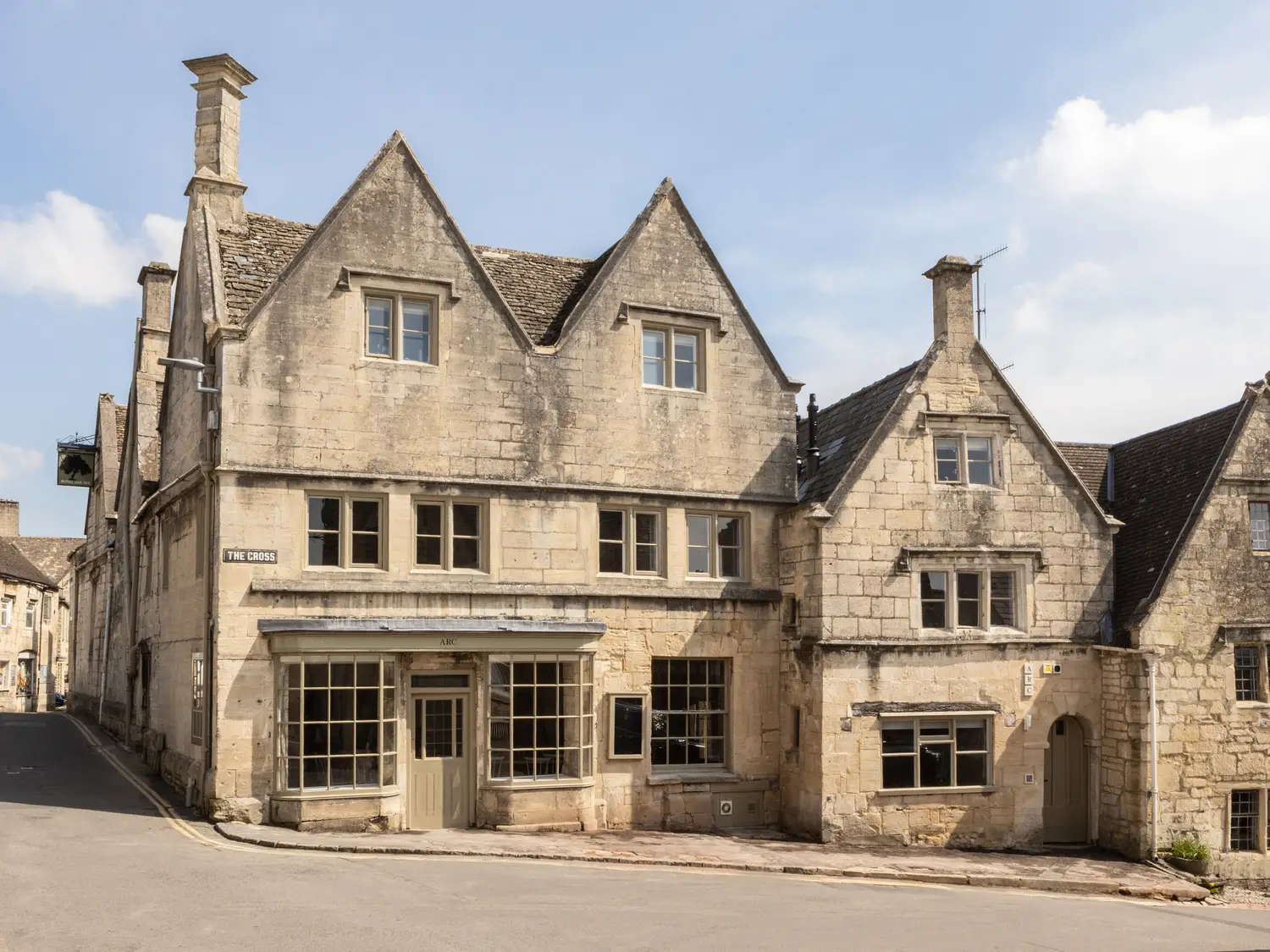 Historic stone building with steep gabled roofs and multiple chimneys, located on a quiet street corner under a blue sky.