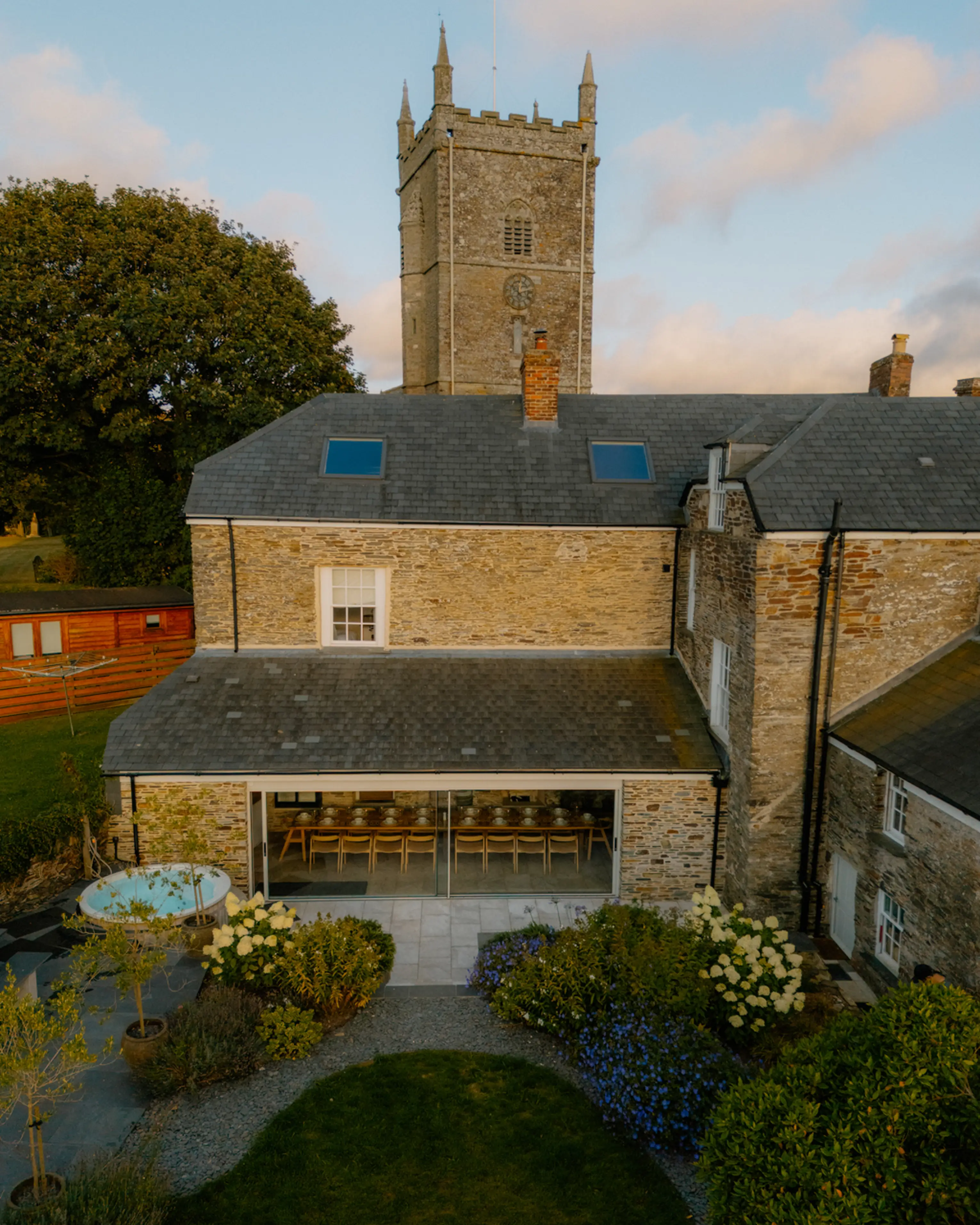 ARC Padstow lit at dusk, the stone manor and converted dining room with the village church tower behind and planted gardens in front