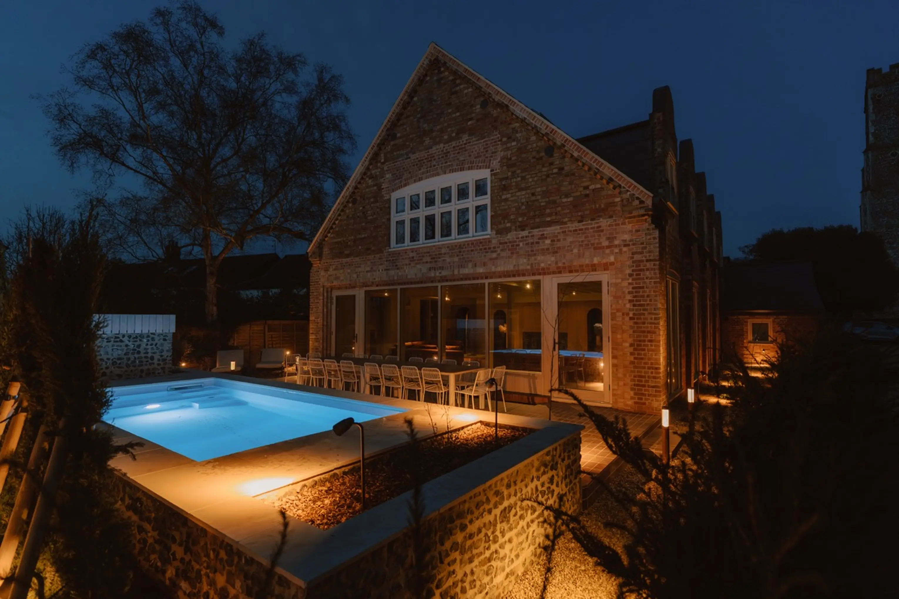 ARC Next-the-Sea at dusk with a lit outdoor pool and the house glowing beyond the garden