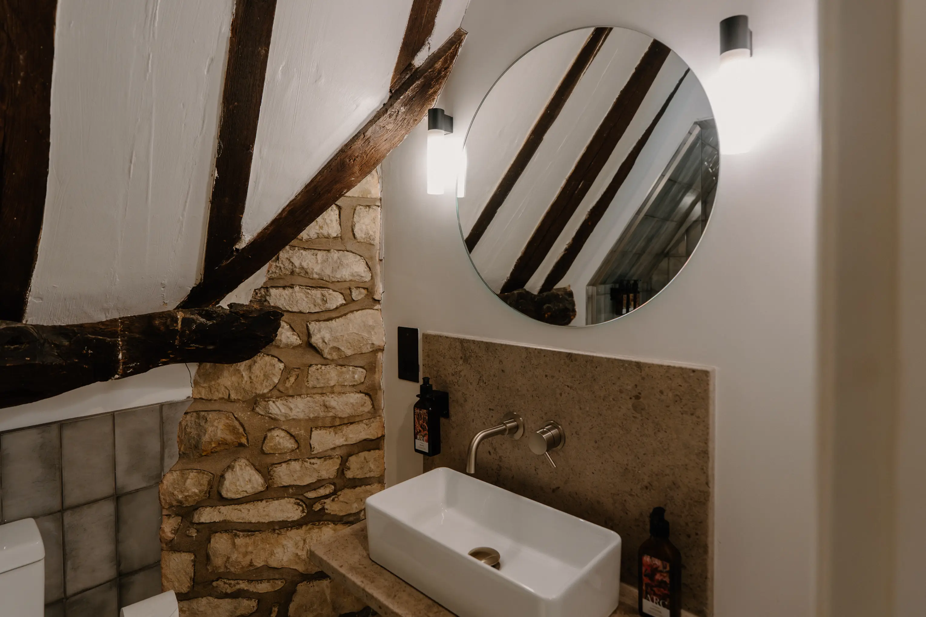 Attic ensuite at ARC Painswick with a round mirror, basin and stone wall under the eaves