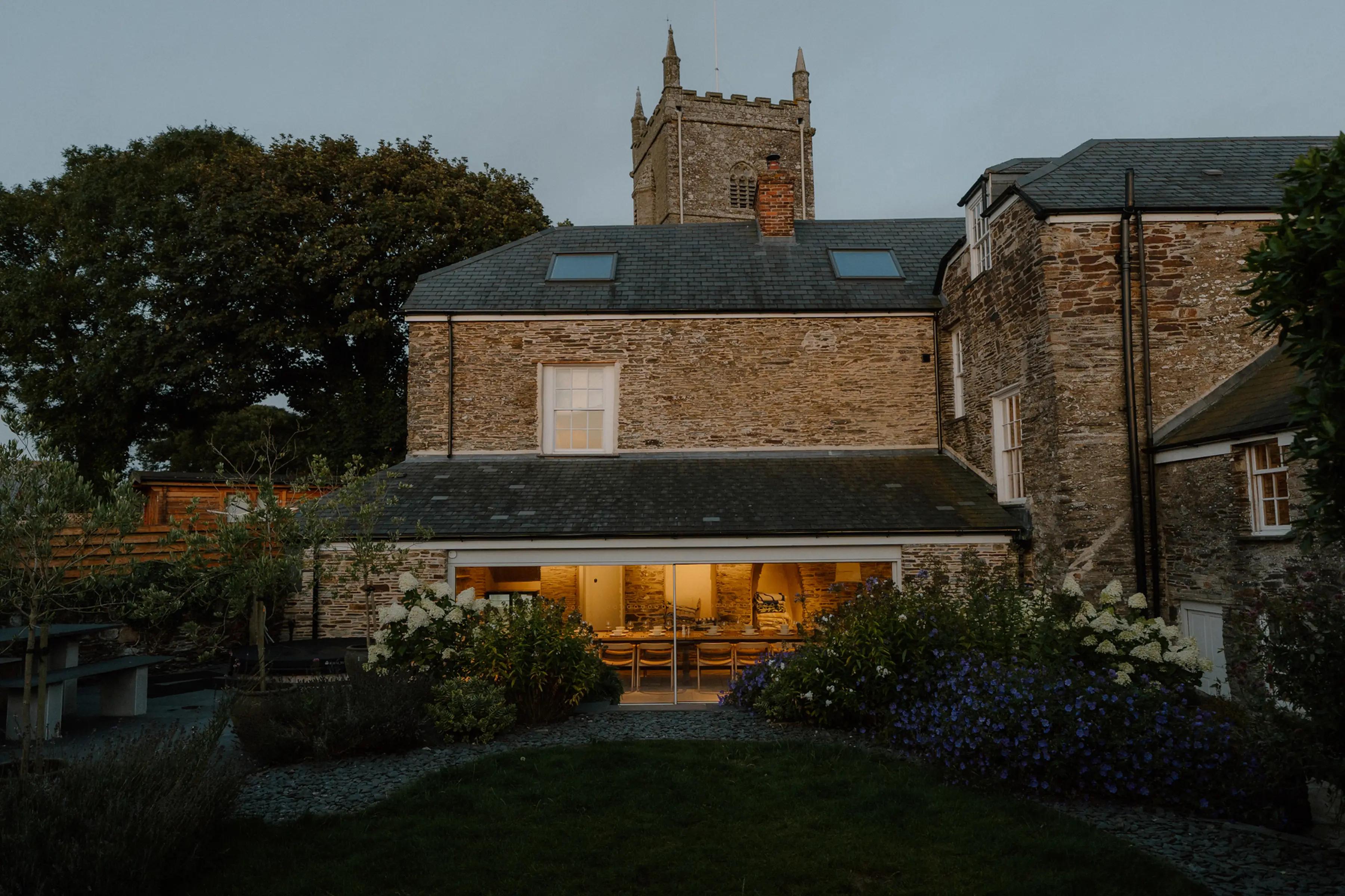 A historic stone house at dusk, warmly lit inside, with a garden in front and a church tower visible in the background.