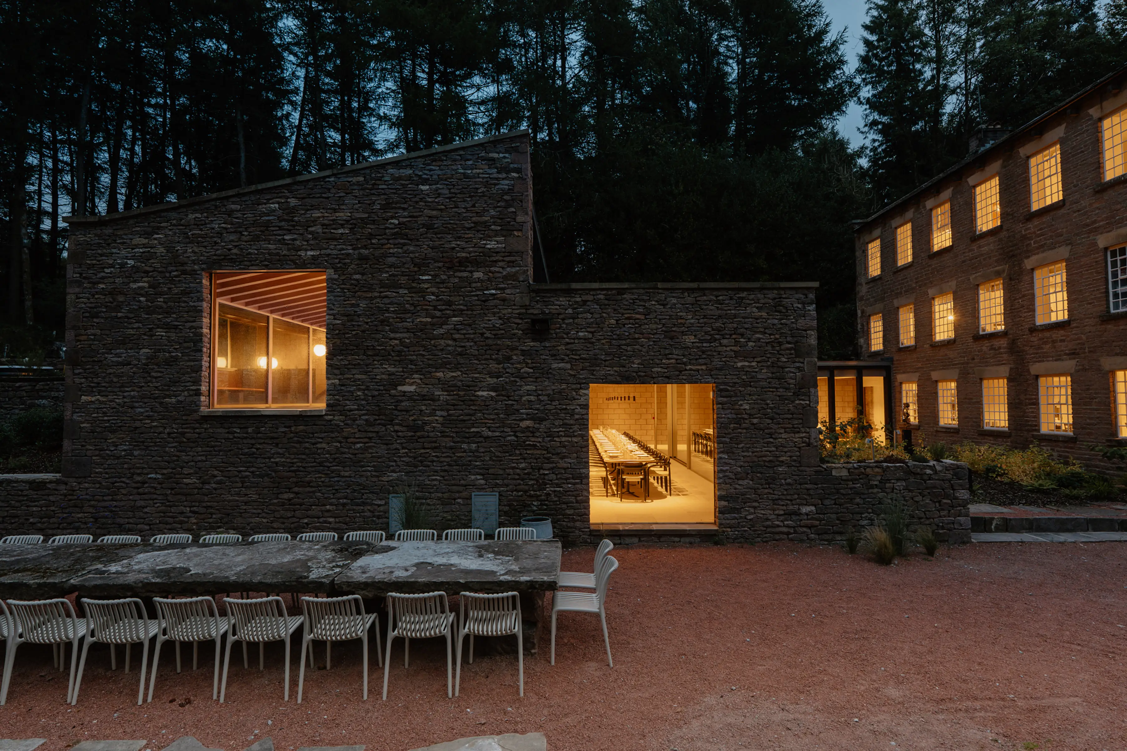ARC Peaks at dusk, a stone former printworks in a Peak District forest clearing with a long outdoor dining table and lit dining hall