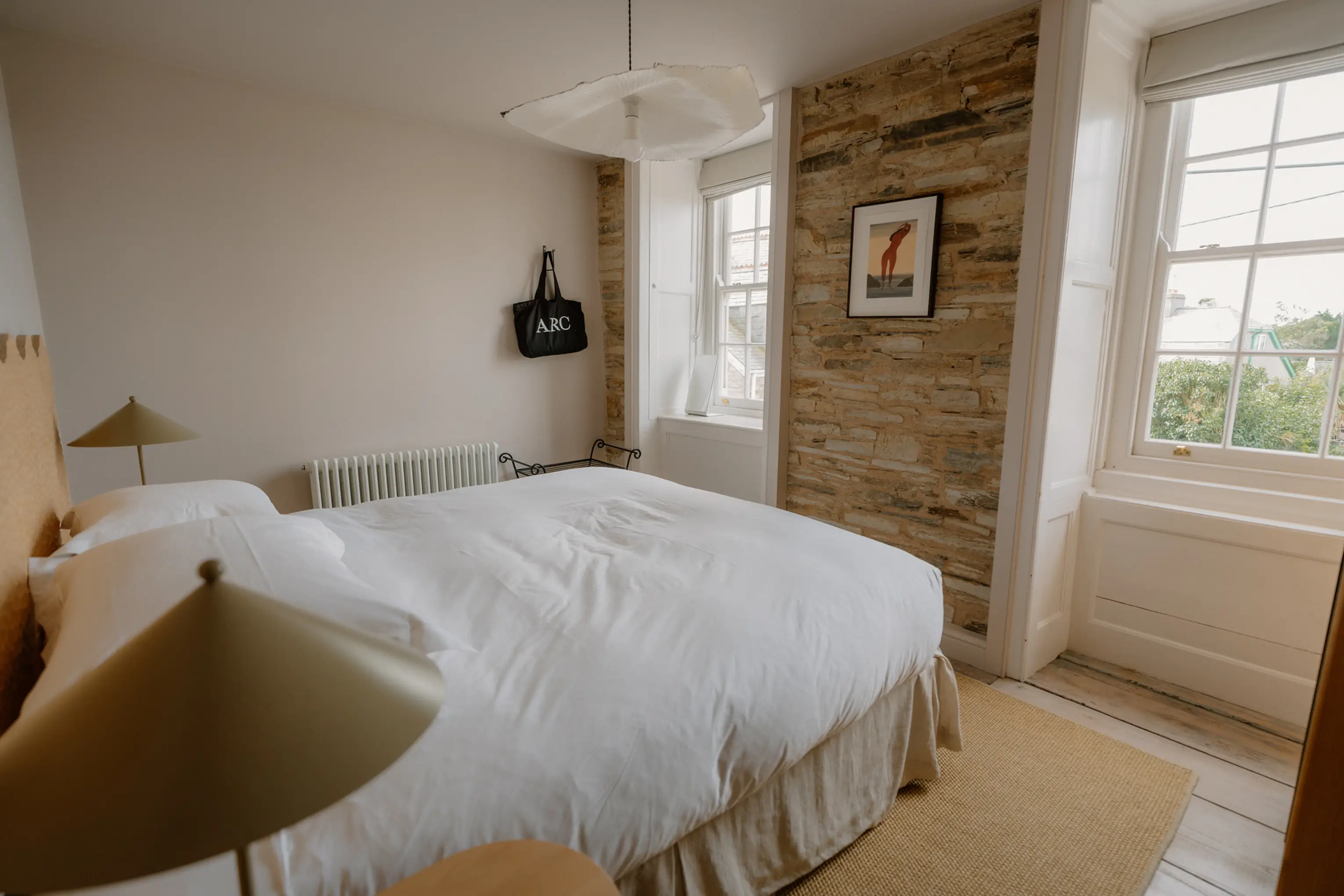Superking bedroom at ARC Padstow with a scalloped timber headboard, an exposed stone wall and two windows