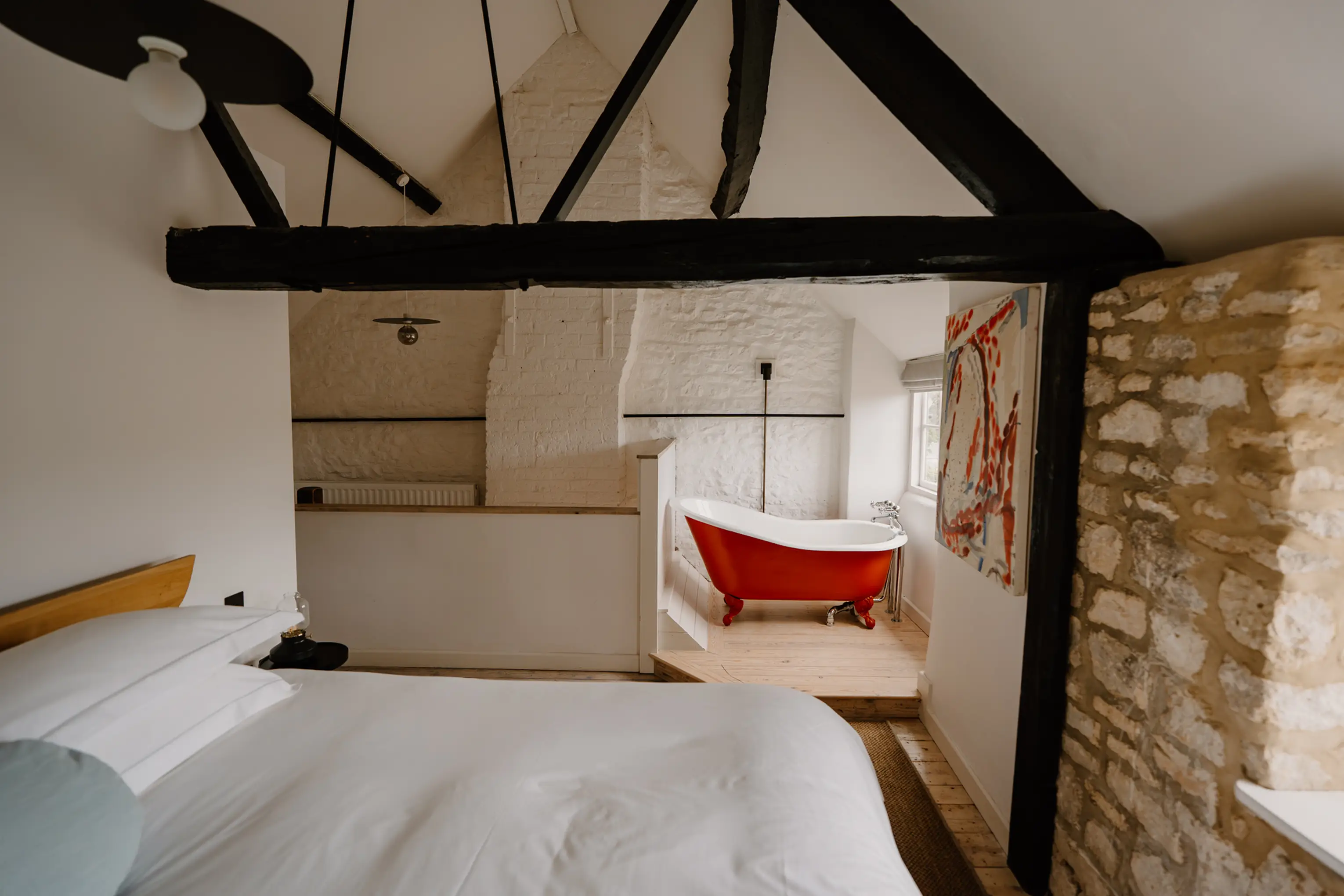 Eaves bedroom at ARC Painswick with exposed beams, a stone wall and a red roll-top bath