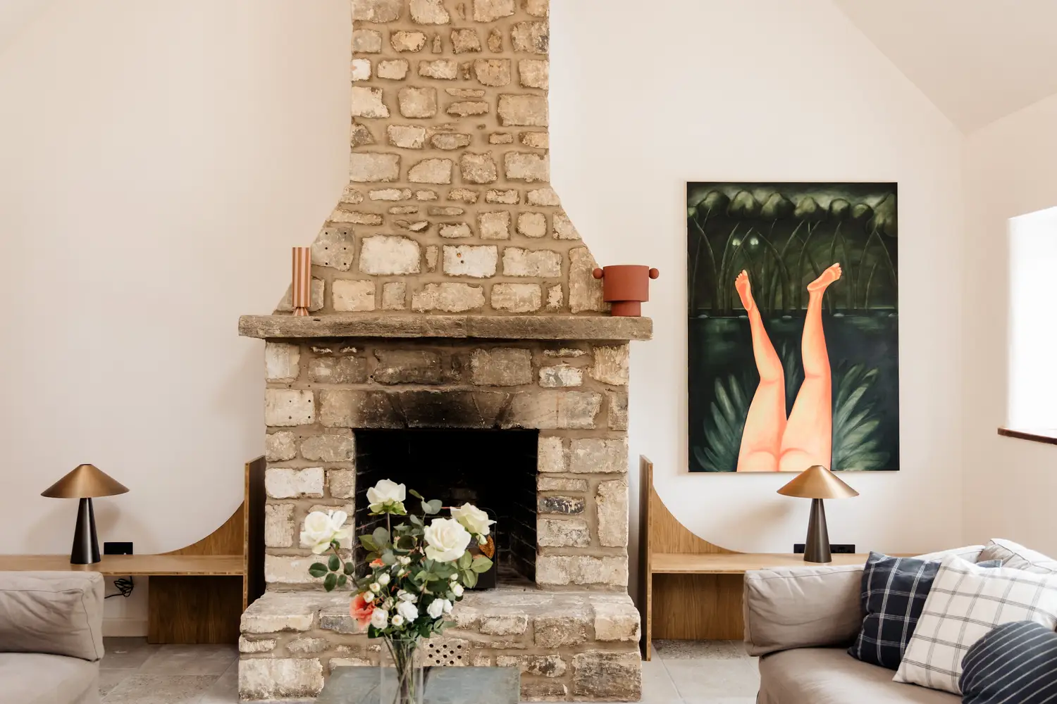 Cozy living room with a stone fireplace, floral arrangement, modern lamps, and artwork of legs in water. Neutral-toned sofas flank the fireplace.