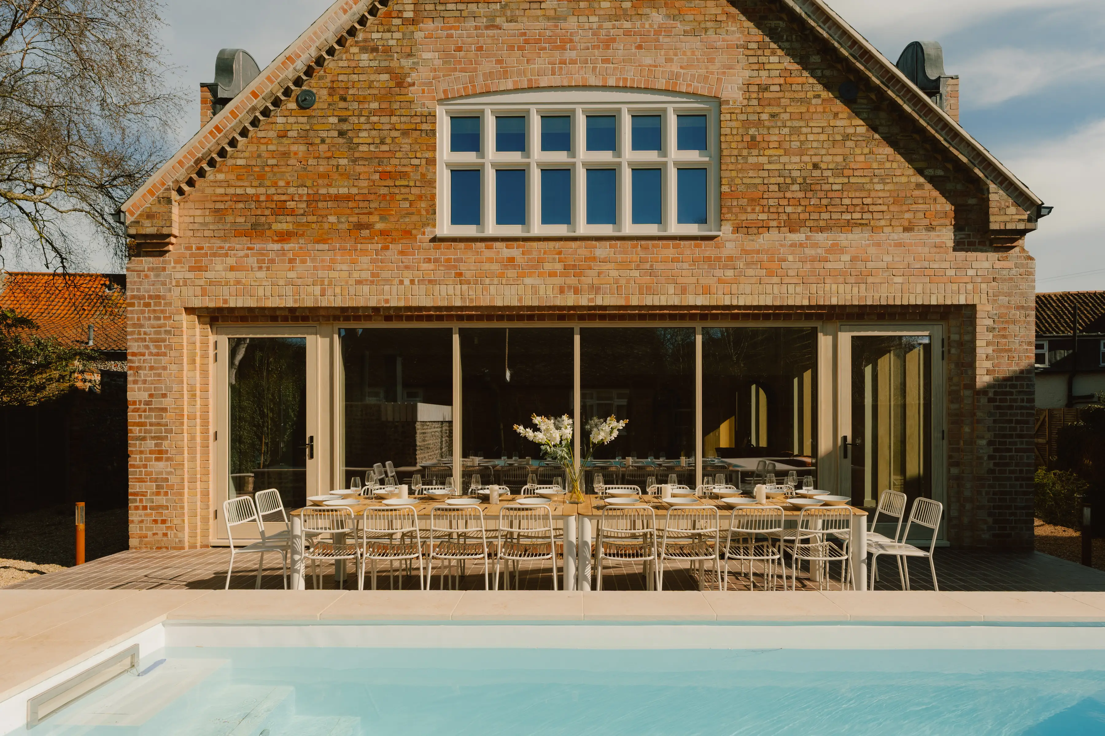 ARC Next-the-Sea in North Norfolk, a brick house with bifold doors open onto a long poolside dining table set for a group