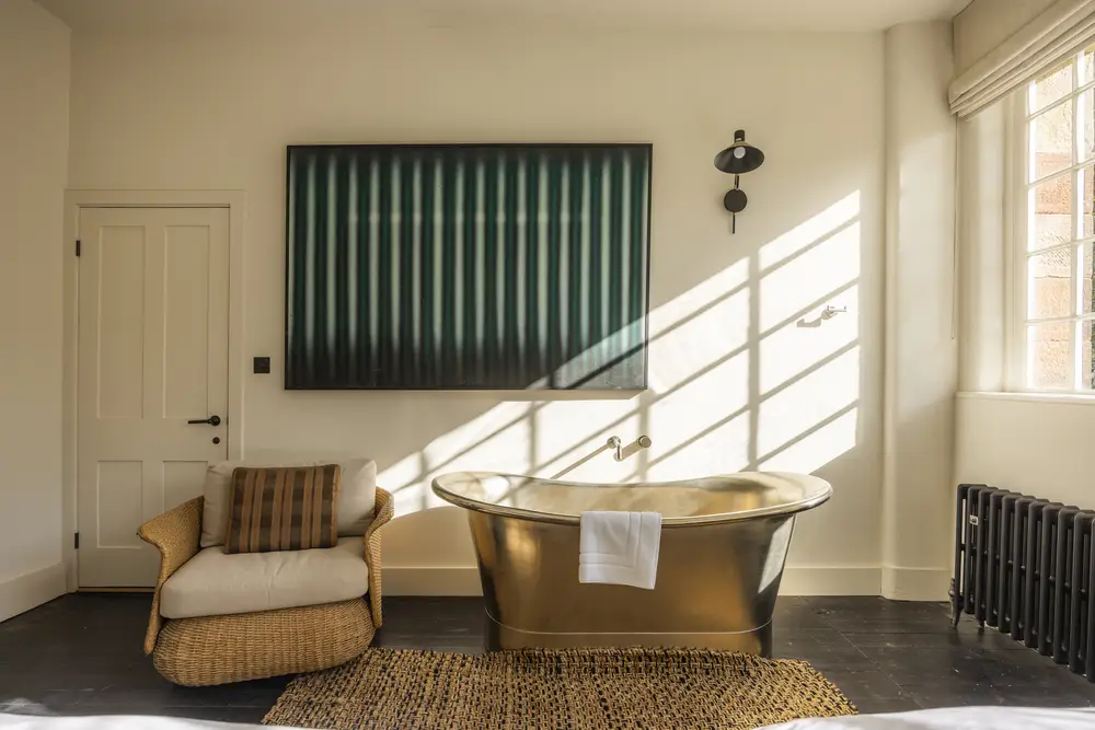 A cozy bathroom in ARC Peak District group accommodation with a brass bathtub, wicker chair, striped artwork, and sunlight streaming through a window onto a textured rug.