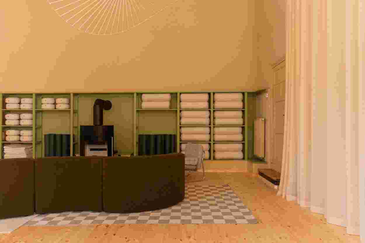 Minimalist room with green shelves holding rolled towels, a black wood stove, curved green sofa, checkered rug, and light wooden floor.