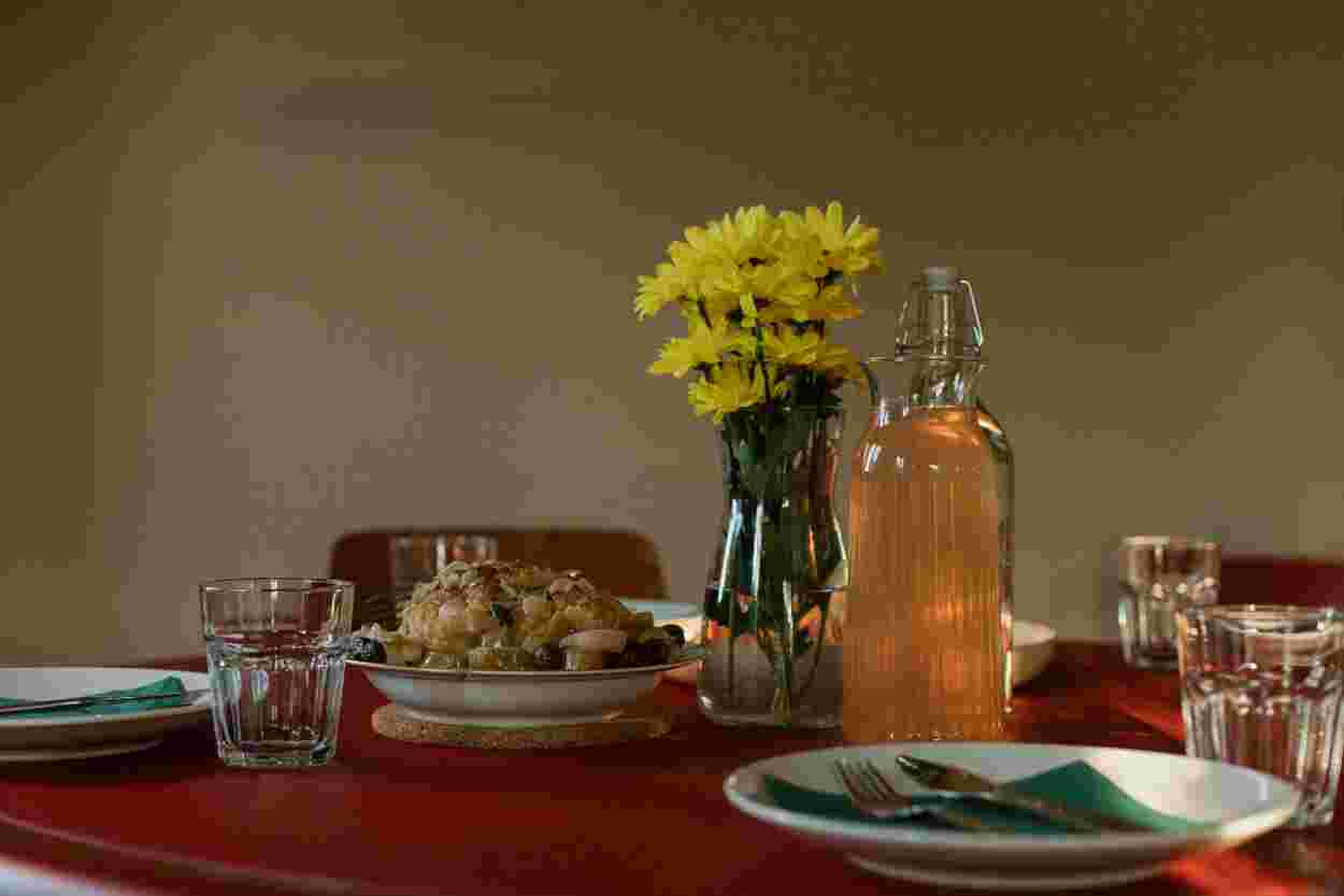 A dining table with a vase of yellow flowers, a glass bottle of juice, plates of food, and glasses on a red tablecloth.