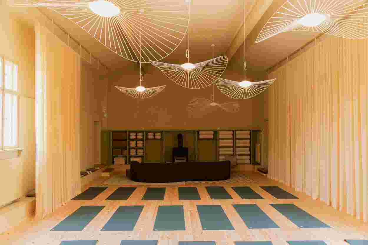 A spacious yoga studio with wooden floors, green mats arranged in rows, soft lighting, and modern, artistic ceiling fixtures.