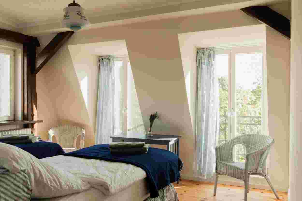 Cozy attic bedroom with two beds, wicker chairs, and a small table. Light blue curtains hang by the windows, letting in natural light.