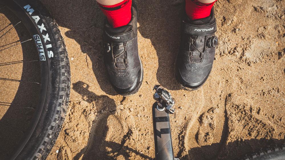 Radschuhe neben einem Mountainbike auf Sand.