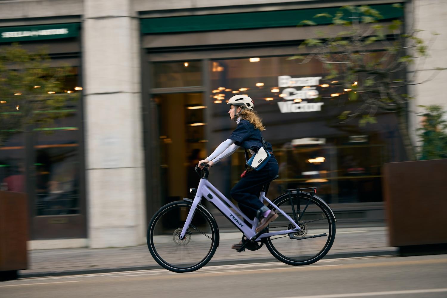 Frau mit Fahrradhelm fährt auf einem hellvioletten Trek Imbound+ durch eine Stadtstraße vor einem Café.