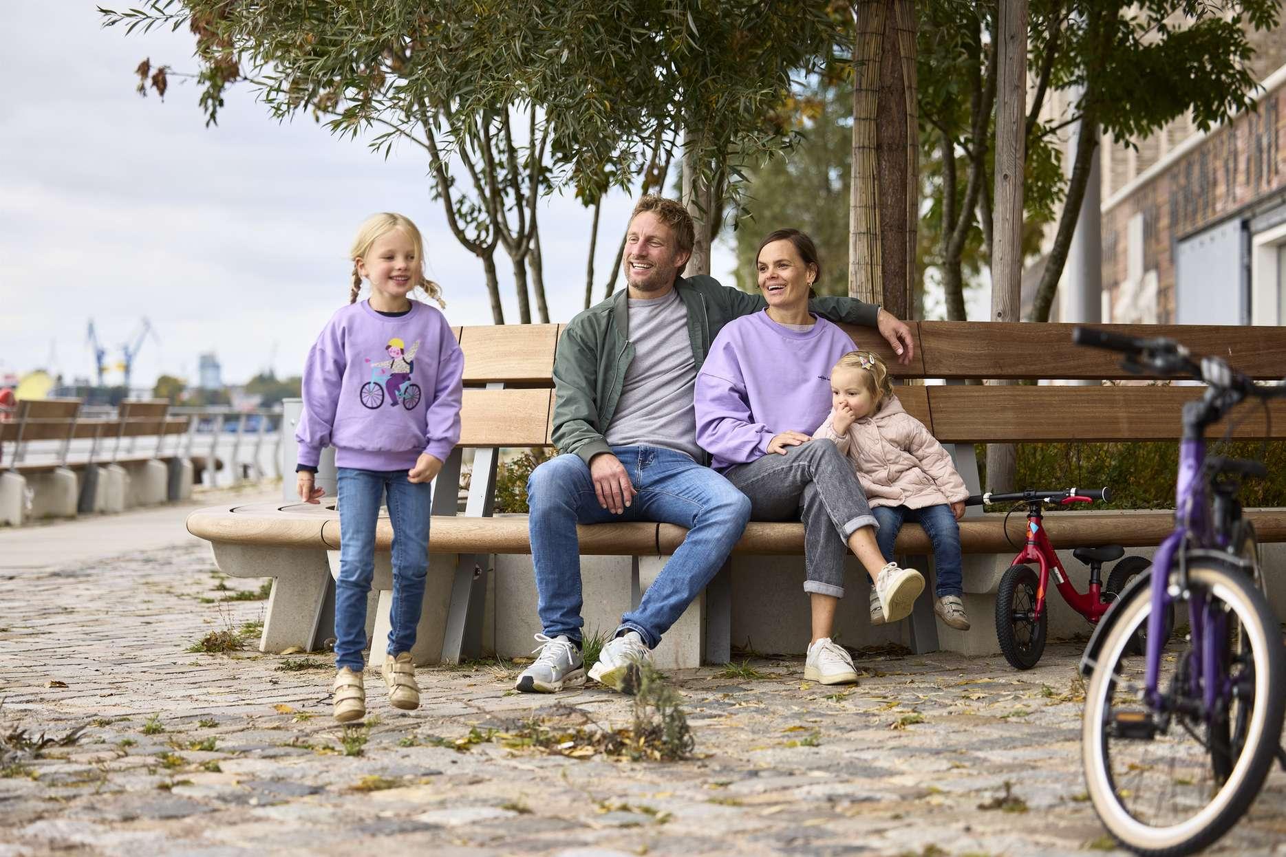 Eine Familie sitzt auf einer Bank am Flussufer, waehrend ein Kind steht und ein Fahrrad daneben geparkt ist.